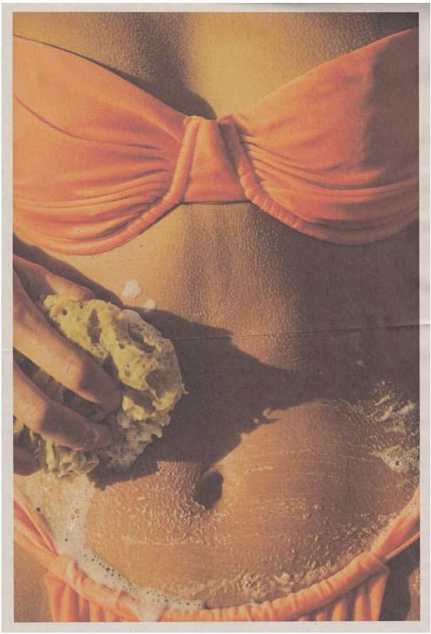 Close-up torso of fair-skinned woman in coral bikini, washing her stomach with a sponge and soapy water in a brightly lit setting.