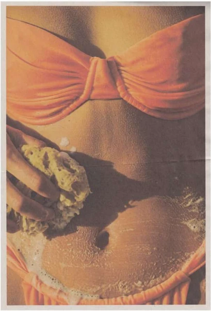 Close-up torso of fair-skinned woman in coral bikini, washing her stomach with a sponge and soapy water in a brightly lit setting.
