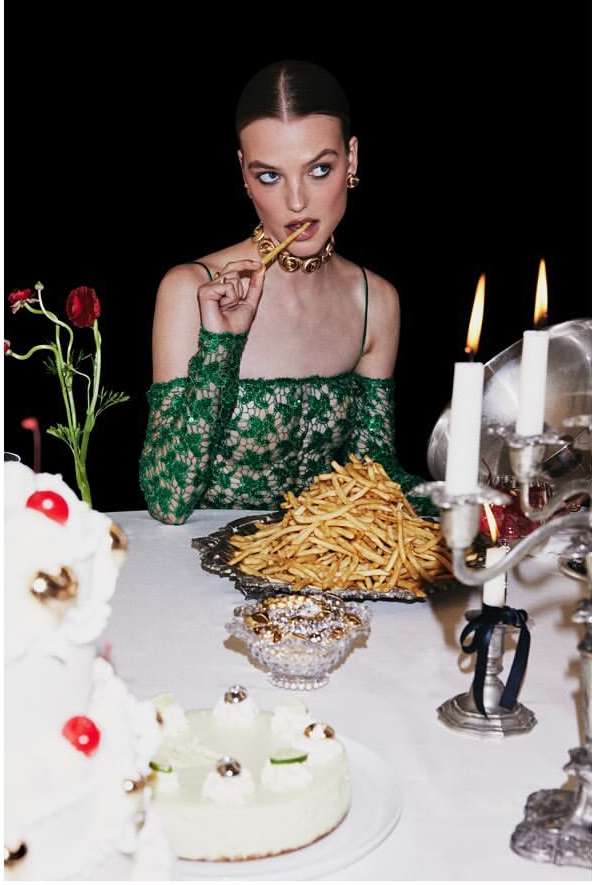 Elegant woman in a green dress eats fries at a table laden with cake and a candelabra against a dark background in a studio shot.