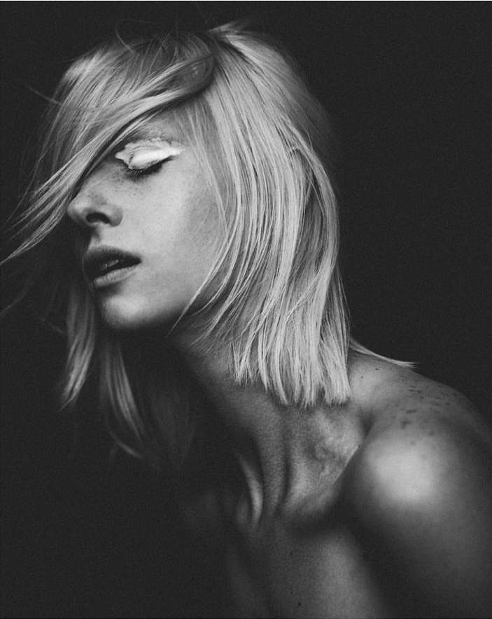 Striking black and white portrait of a woman with blonde hair and white makeup around her eyes, with a dark background.