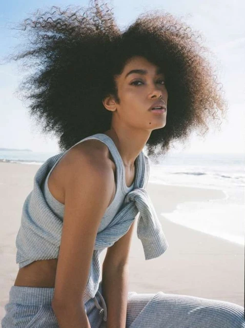 Woman with voluminous afro hairstyle sitting on a sandy beach in casual clothing, with ocean water and sky visible in the background.