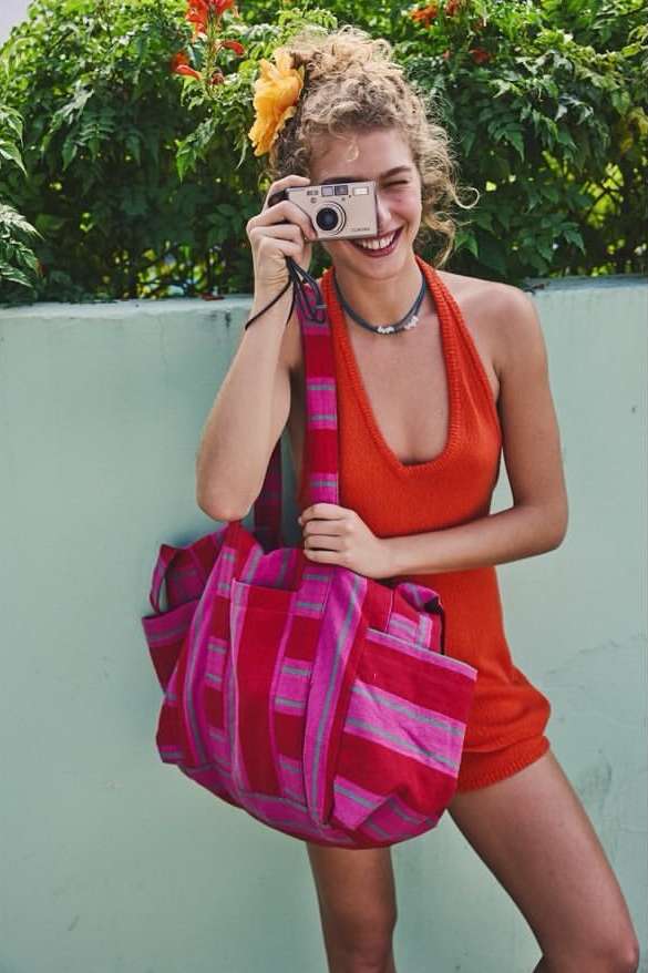 A young woman smiles and holds a camera to her eye. She carries a colorful striped tote bag, stands outdoors against green foliage.
