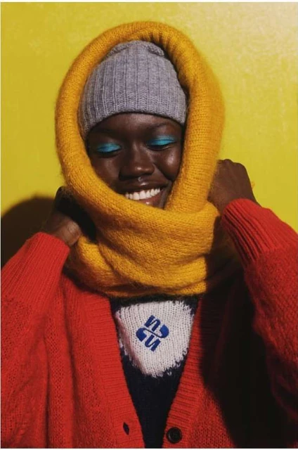 A smiling Black model in red cardigan and gray beanie hat poses against a yellow background, framed by a gold-colored knit scarf.