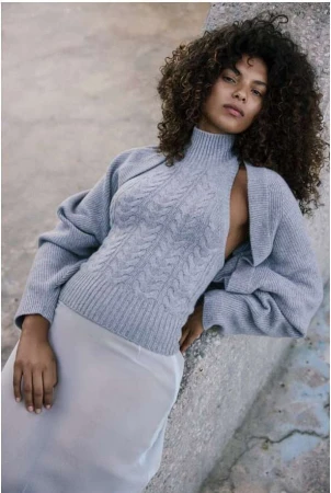 Woman with voluminous curly hair leaning against a stone wall wearing a grey turtleneck knit sweater and light skirt.