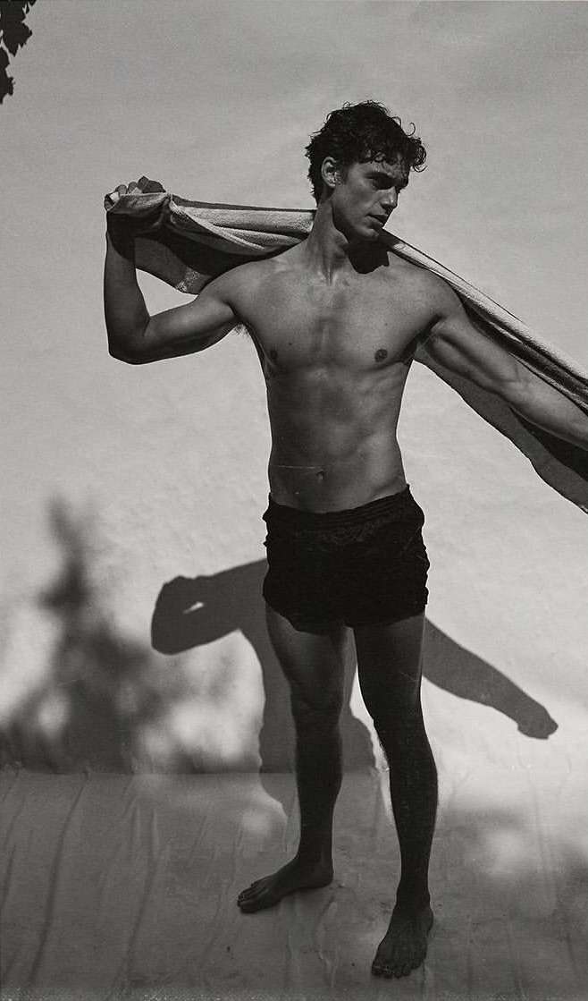 Monochrome portrait of a shirtless adult male with dark hair, black shorts, and a towel draped over his shoulder in an outdoor setting.
