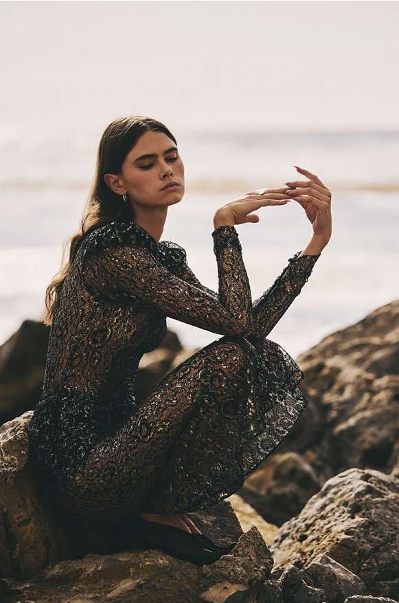 A woman in a long, dark lace dress crouches on rocks near the beach, with the sea and sky visible in the background, captured in a fashion-style photograph.