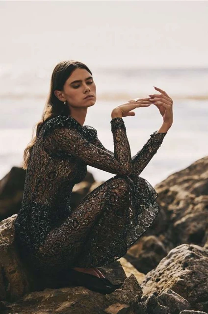A woman in a long, dark lace dress crouches on rocks near the beach, with the sea and sky visible in the background, captured in a fashion-style photograph.