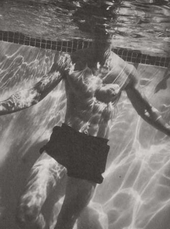 Monochrome underwater photograph of a shirtless man swimming in a pool with rippled light patterns reflecting across his body.