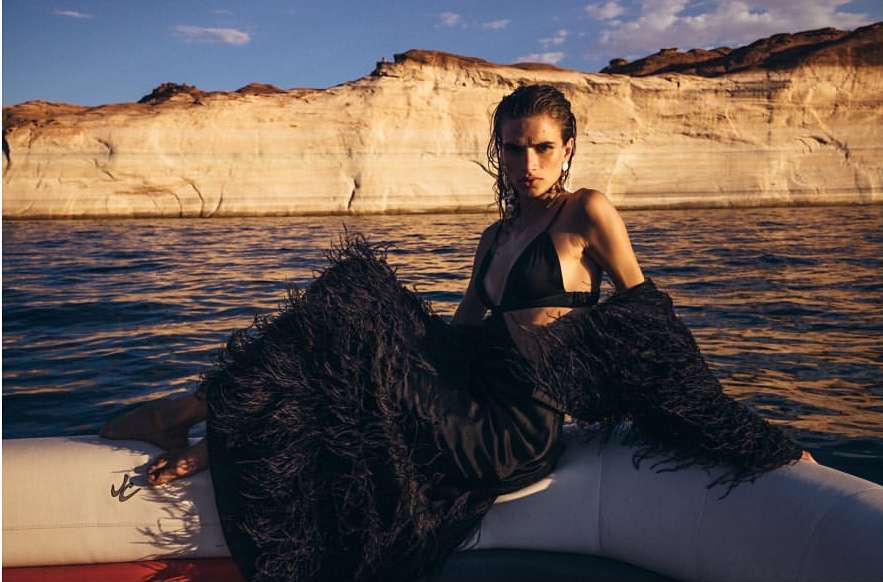 A young woman with dark hair sits on an inflatable boat on a lake with light reflecting on the water in front of rock formations.