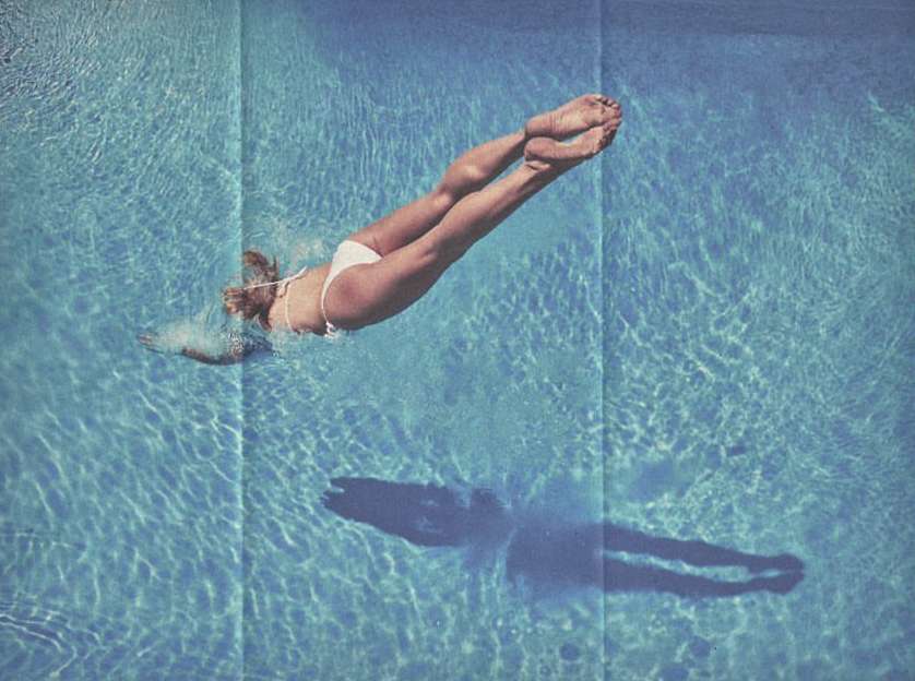 Aerial view of a woman in a white bikini diving gracefully into a turquoise blue swimming pool with sunlight creating water patterns.