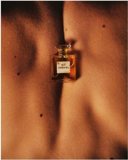 Close-up shot of a Chanel No. 5 perfume bottle placed between two torsos, bathed in warm light highlighting skin textures.