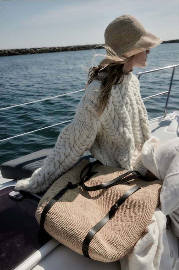 A woman wearing a white cable knit sweater and sun hat is sitting on a boat with a large woven bag, ocean and blue sky in the background.