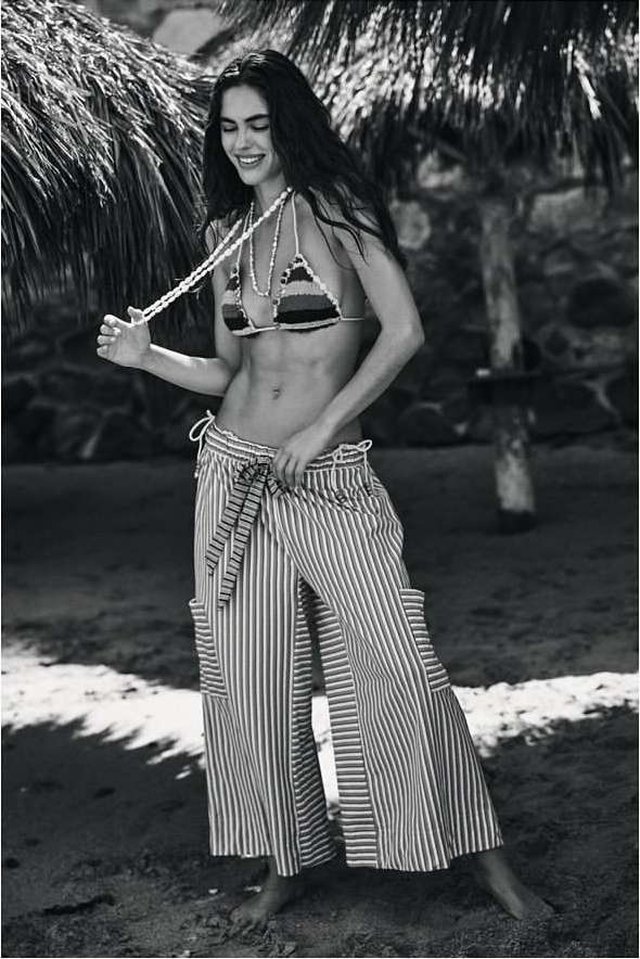 Monochrome portrait of a smiling woman in a striped bikini top and trousers standing beneath palm trees.