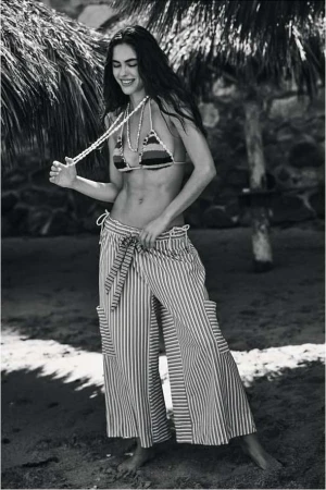 Monochrome portrait of a smiling woman in a striped bikini top and trousers standing beneath palm trees.