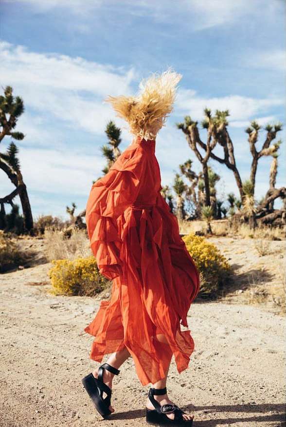 Woman in a red dress walks through a desert landscape with sparse vegetation and Joshua Trees under a partly cloudy blue sky.