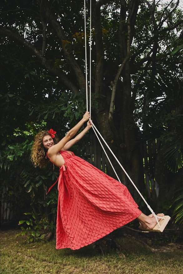 A smiling woman in a red dress swinging on a rope swing in a yard with a large tree providing shade and a tropical vibe.