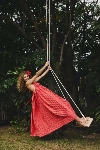 A smiling woman in a red dress swinging on a rope swing in a yard with a large tree providing shade and a tropical vibe.