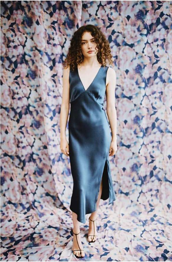 Woman with curly hair in a blue sleeveless dress standing in front of a floral backdrop, facing forward in a fashion pose.
