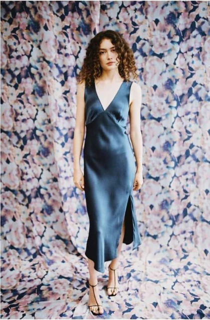 Woman with curly hair in a blue sleeveless dress standing in front of a floral backdrop, facing forward in a fashion pose.