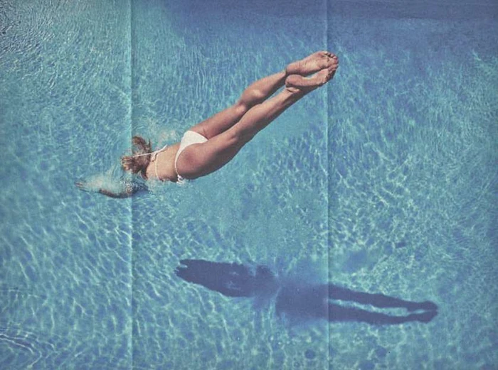 High-angle shot of a woman diving into an outdoor swimming pool, casting a dark shadow on the water surface.