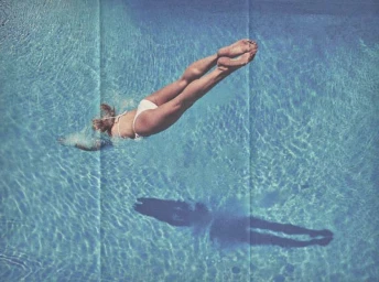High-angle shot of a woman diving into an outdoor swimming pool, casting a dark shadow on the water surface.