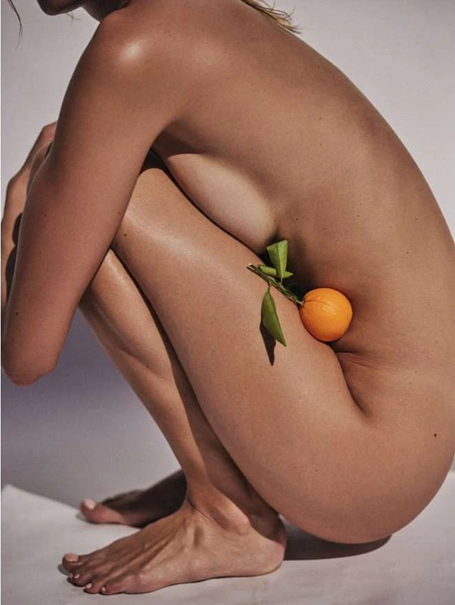 Crouching nude model holding an orange with green leaves against a neutral white background