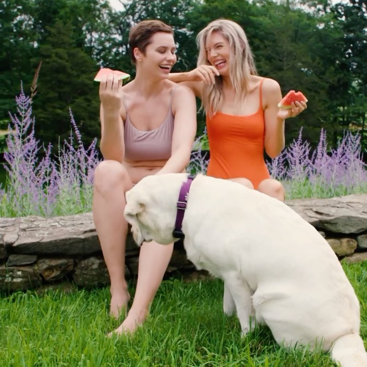Two women in swimwear share watermelon with a dog on a sunny day outdoors, sitting on a stone wall in a grassy area near flowers and trees.