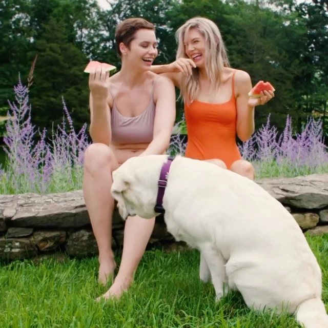 Two women in swimwear share watermelon with a dog on a sunny day outdoors, sitting on a stone wall in a grassy area near flowers and trees.
