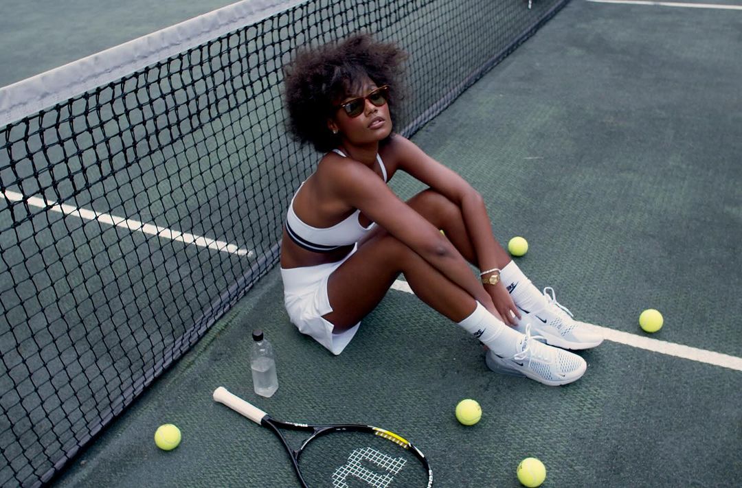 A woman with sunglasses in white tennis clothes sits on a tennis court next to a net with a racquet and tennis balls scattered around.