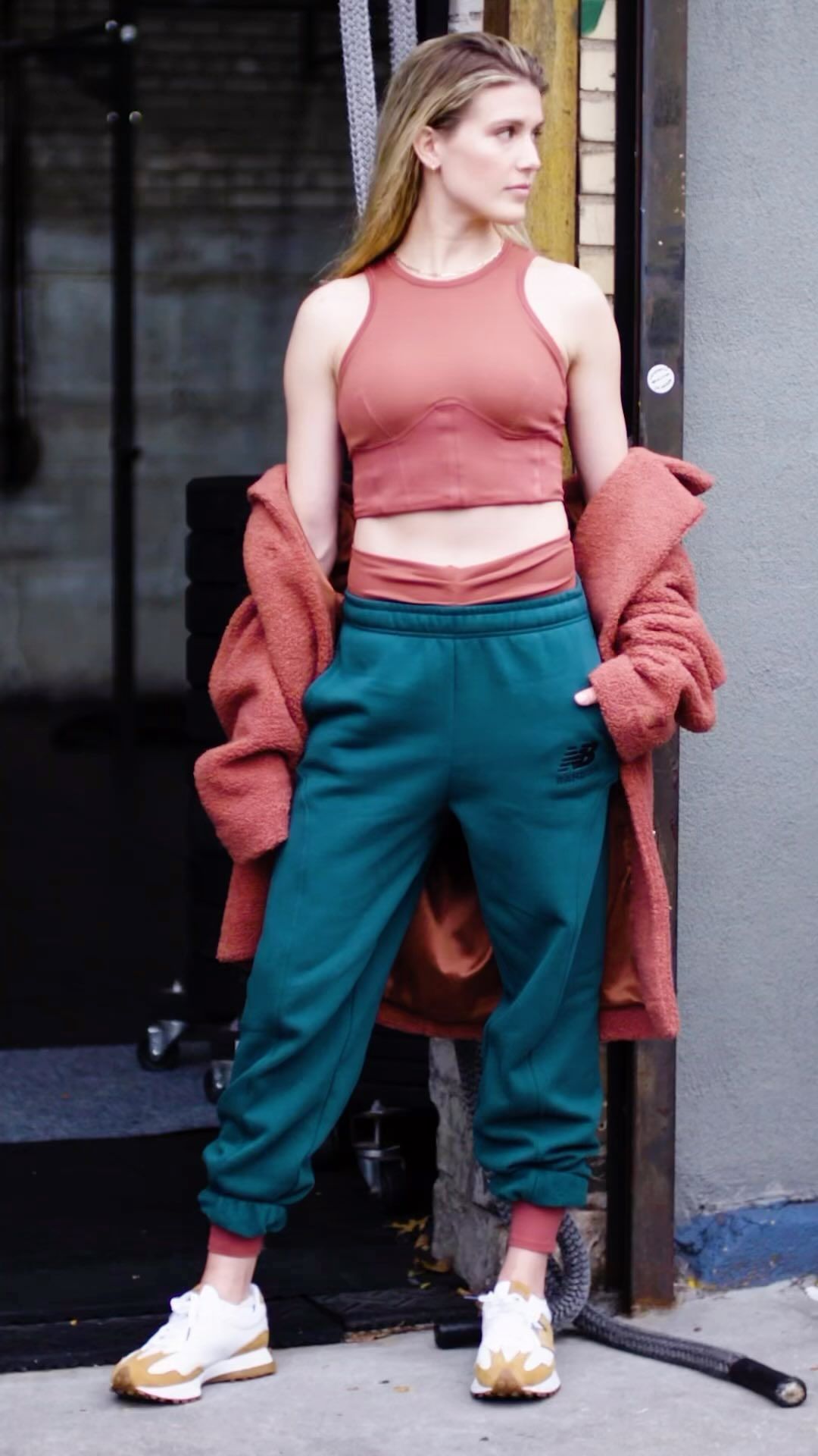 Athletic woman in workout attire posing in an urban setting. She wears a crop top, sweatpants, jacket, and sneakers. 