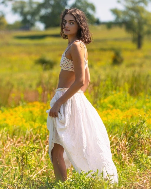 Woman poses in a floral bikini top and white skirt in a sunlit field with yellow flowers and tall grass, under a blurred sky.