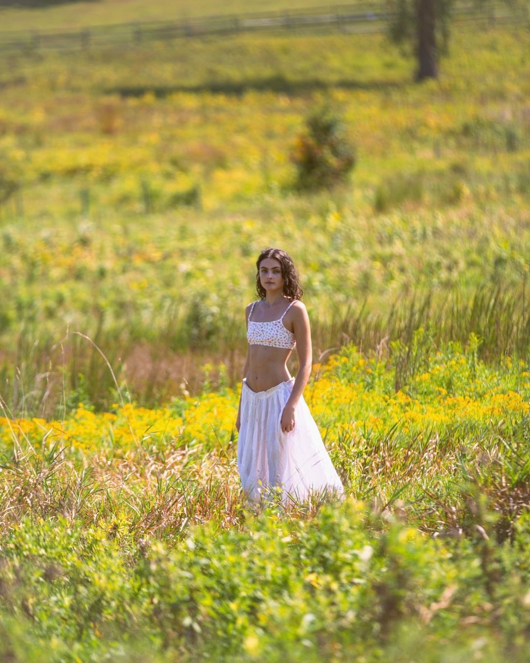 A young woman standing in a lush grassy meadow filled with yellow wildflowers, wearing a white crop top and flowing white skirt.