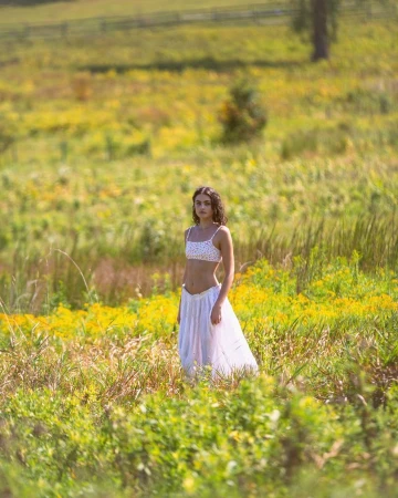 A young woman standing in a lush grassy meadow filled with yellow wildflowers, wearing a white crop top and flowing white skirt.