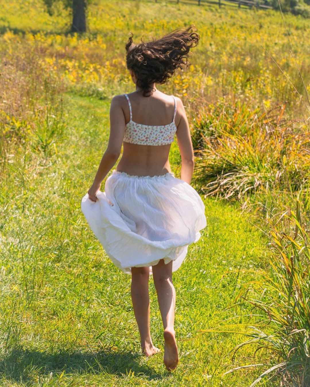 An adult woman with curly hair runs barefoot through a meadow with a white skirt and floral top on a sunny day.