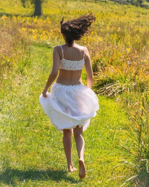 An adult woman with curly hair runs barefoot through a meadow with a white skirt and floral top on a sunny day.