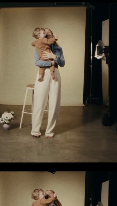 A fair-skinned woman in denim holds a toddler with blonde hair against a neutral backdrop, with a stool, flowers, and lighting visible.