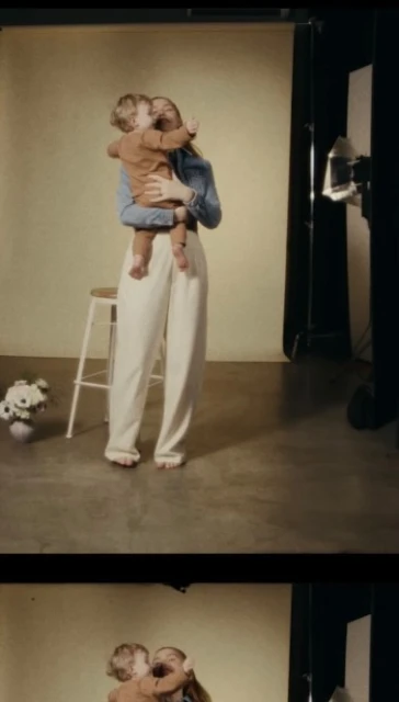 A fair-skinned woman in denim holds a toddler with blonde hair against a neutral backdrop, with a stool, flowers, and lighting visible.