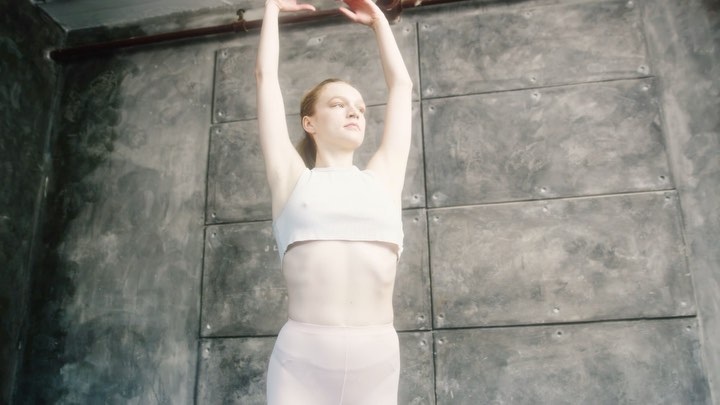 Young woman in a light top and pants poses with arms raised in front of a mottled gray wall in an indoor setting with diffused lighting.