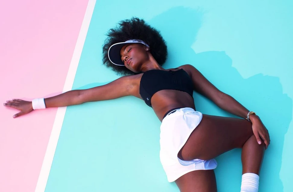 An African-American woman in athletic wear poses against a split pink and teal background, stretching her arm and leg, wearing a visor and wristband.