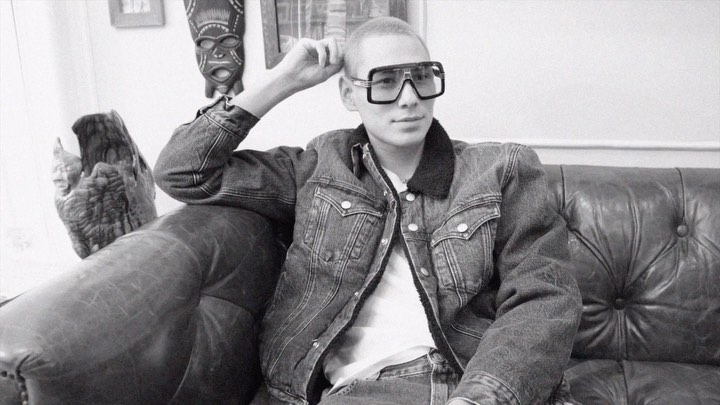 Black and white portrait of a young adult in sunglasses and denim jacket, seated on a vintage leather sofa, with art objects in the background.
