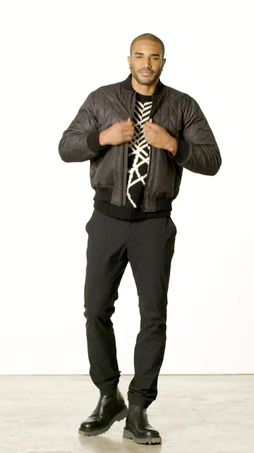 Full shot of a light-skinned adult man in a bomber jacket, patterned sweater, pants, and boots standing in a photo studio against a white backdrop.