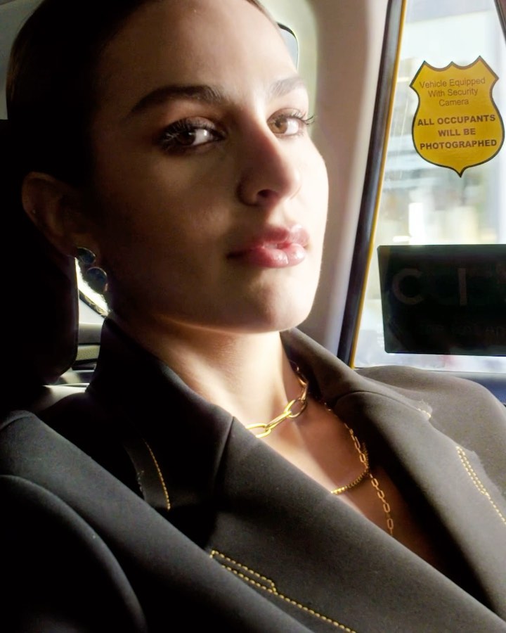 Attractive woman in a black blazer and gold necklaces, sitting in the back of a taxi, looking at the camera with a neutral expression.