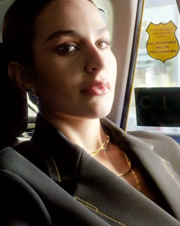 Attractive woman in a black blazer and gold necklaces, sitting in the back of a taxi, looking at the camera with a neutral expression.