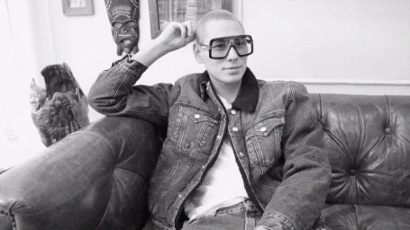 Monochrome portrait of a man wearing oversized glasses and a denim jacket seated on a leather sofa