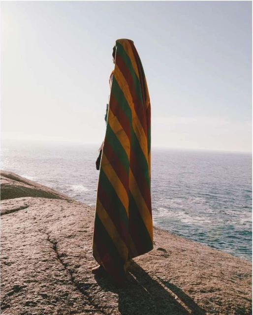 Person draped in a striped textile on a rocky outcrop, overlooking ocean and clear sky. Warm, natural lighting highlights the textured fabric.