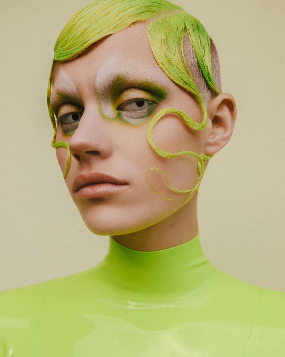 Close-up portrait of a person with vibrant green and white makeup, short green hair, and a high-necked green shirt against a yellow backdrop.