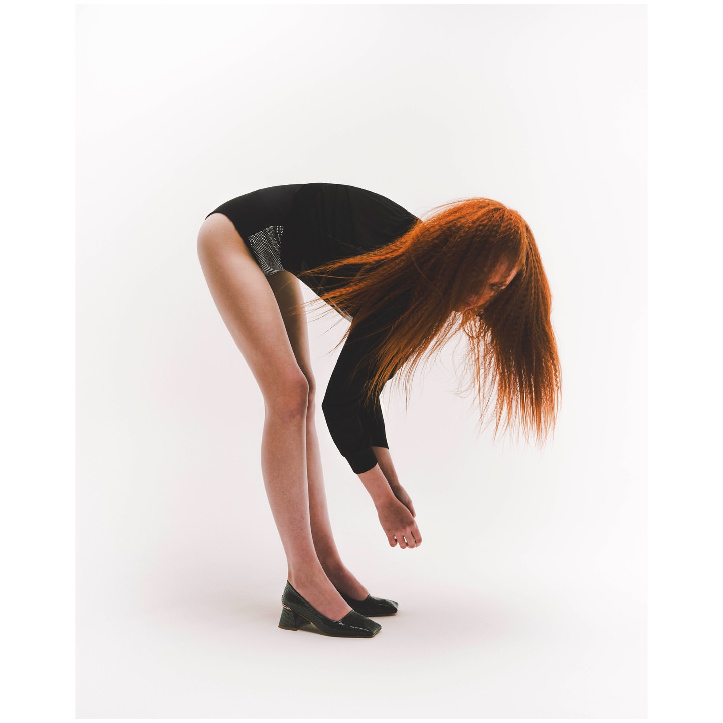 Woman in a black leotard bends over in front of a white backdrop in a studio setting. Her red hair hangs down toward the floor.