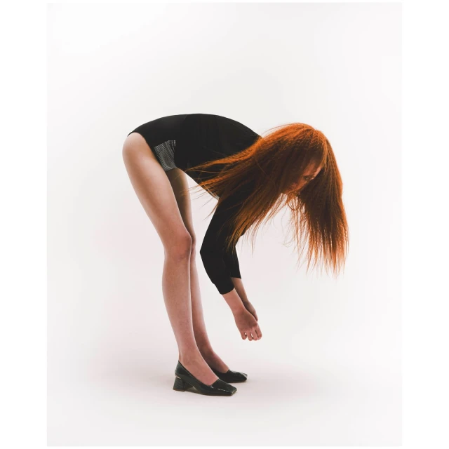 Woman in a black leotard bends over in front of a white backdrop in a studio setting. Her red hair hangs down toward the floor.