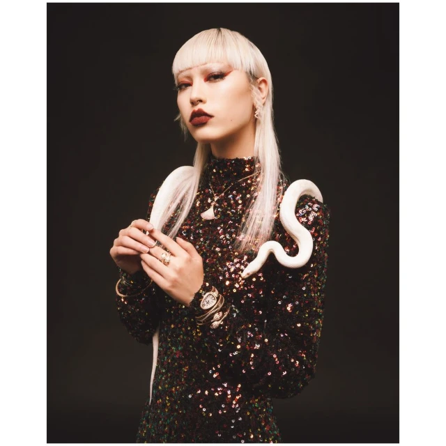 Woman with short bleached hair poses in a sequin dress with an ivory snake draped over her shoulder against a dark background.