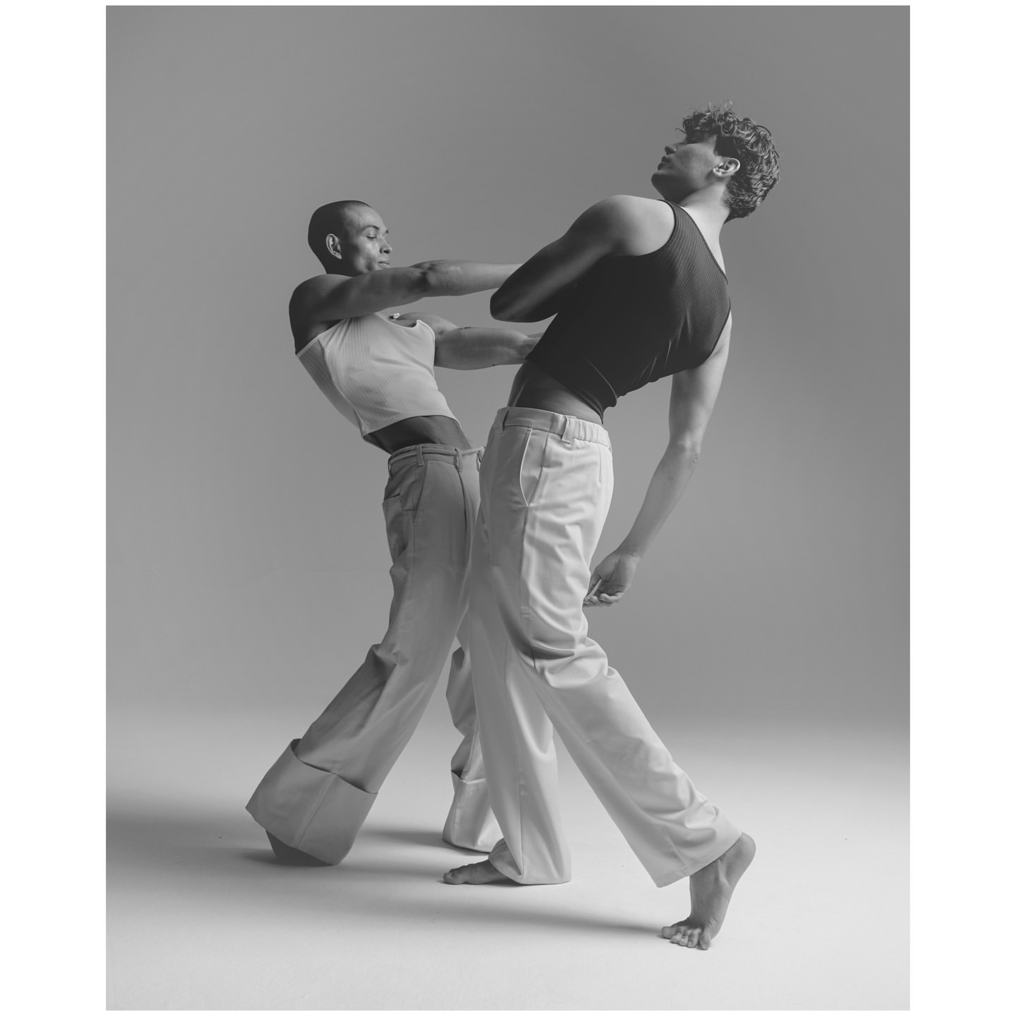Monochromatic shot of two slender figures in a dynamic pose wearing relaxed trousers and tank tops, set against a simple studio backdrop.